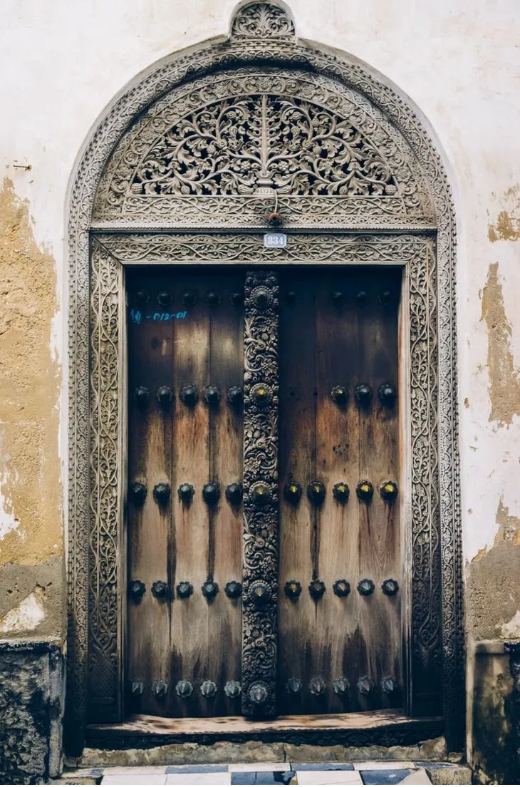A weathered carved door in Stone Town, Zanzibar, its timber leaves studded with brass bosses beneath an intricately carved arched lintel