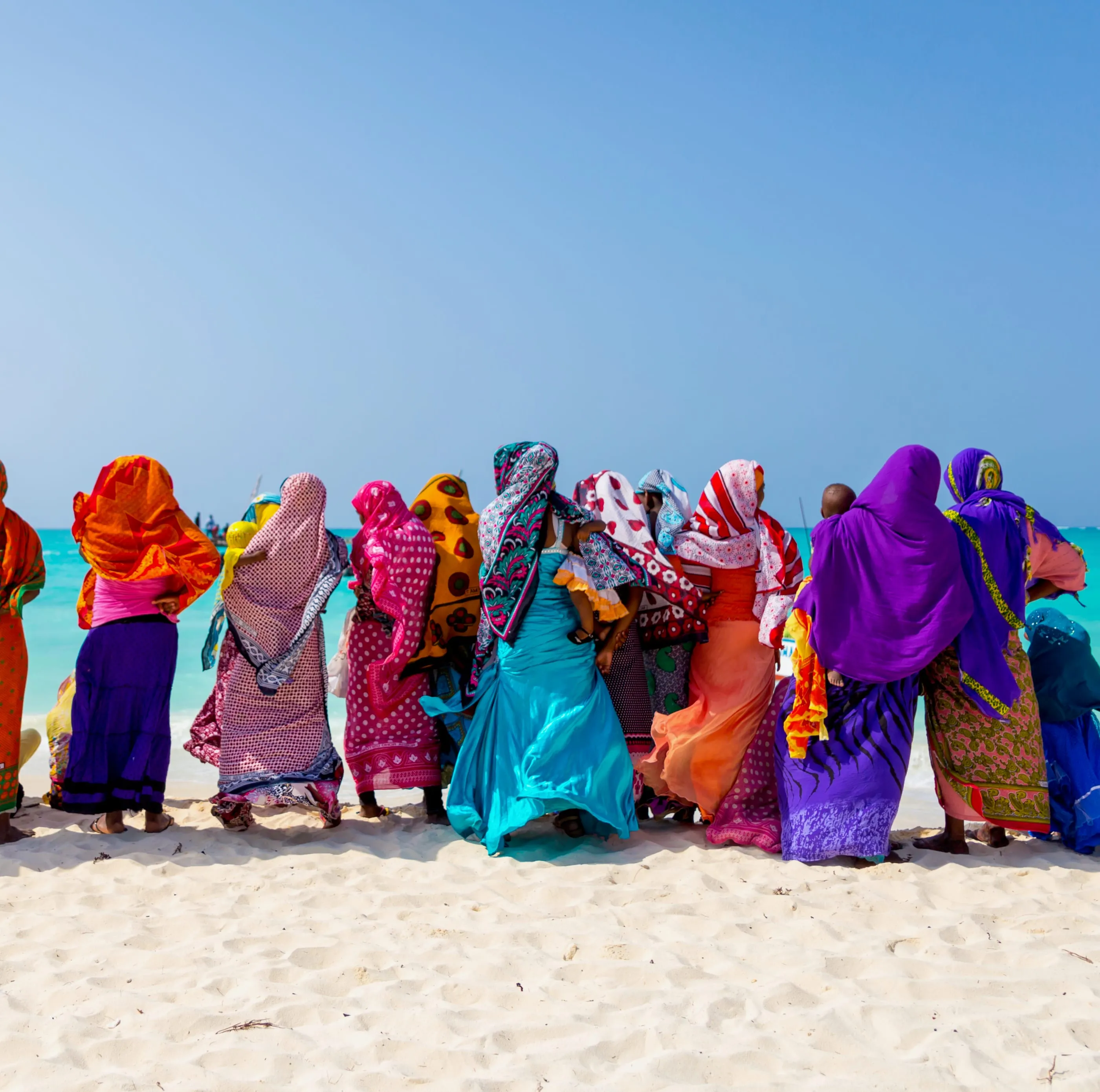 Women wrapped in brightly patterned kangas stand in a line on a white-sand beach, looking out to sea in Zanzibar